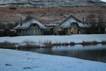 Two thatched-roof cottages are situated in a snowy landscape beside a calm body of water. The snow covers the ground and rooftops, creating a soft and serene atmosphere. The surrounding hills are lightly dusted with snow, and some trees can be seen in the background. The warm light from the windows suggests a cozy interior.