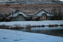 Two thatched-roof cottages are situated in a snowy landscape beside a calm body of water. The snow covers the ground and rooftops, creating a soft and serene atmosphere. The surrounding hills are lightly dusted with snow, and some trees can be seen in the background. The warm light from the windows suggests a cozy interior.
