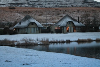 Two thatched-roof cottages are situated in a snowy landscape beside a calm body of water. The snow covers the ground and rooftops, creating a soft and serene atmosphere. The surrounding hills are lightly dusted with snow, and some trees can be seen in the background. The warm light from the windows suggests a cozy interior.