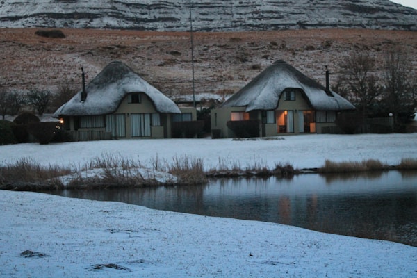 Two thatched-roof cottages are situated in a snowy landscape beside a calm body of water. The snow covers the ground and rooftops, creating a soft and serene atmosphere. The surrounding hills are lightly dusted with snow, and some trees can be seen in the background. The warm light from the windows suggests a cozy interior.