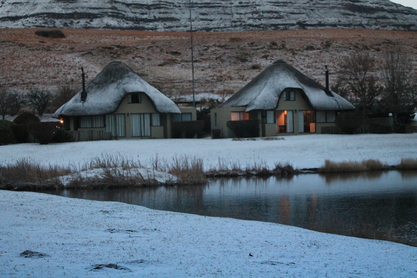 Two thatched-roof cottages are situated in a snowy landscape beside a calm body of water. The snow covers the ground and rooftops, creating a soft and serene atmosphere. The surrounding hills are lightly dusted with snow, and some trees can be seen in the background. The warm light from the windows suggests a cozy interior.