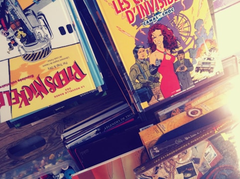 A collection of comic books is displayed, with covers featuring colorful illustrations. One prominent cover shows a woman in a red dress, accompanied by various characters and an amusement park ride in the background. Another comic cover displays a dynamic scene with a construction vehicle and workers.