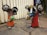 Two whimsical sculptures resembling mice are crafted from scrap metal and painted in bright colors. They have exaggerated features like large ears and eyes made from recycled parts. The sculptures are standing on a stone pavement, and there is a sign in the background indicating a sale.