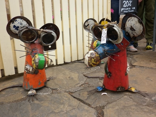 Two whimsical sculptures resembling mice are crafted from scrap metal and painted in bright colors. They have exaggerated features like large ears and eyes made from recycled parts. The sculptures are standing on a stone pavement, and there is a sign in the background indicating a sale.