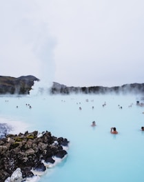 People enjoy bathing in a natural geothermal hot spring surrounded by rocks. The water is a striking blue-green color, and steam rises into the overcast sky. Hills and sparse vegetation are visible in the background.