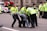 Police officers in bright yellow jackets are carrying a person away from the scene. The person is wearing a dark jacket and jeans. In the background, there are more police officers and a police motorcycle. A red van and buildings are visible in the background.