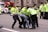 Police officers in bright yellow jackets are carrying a person away from the scene. The person is wearing a dark jacket and jeans. In the background, there are more police officers and a police motorcycle. A red van and buildings are visible in the background.