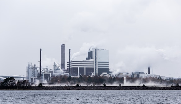 A large industrial facility emits smoke and steam into a cloudy sky. The complex appears to be near a body of water, with visible chimneys and various structures. The surrounding area includes sparse trees and a concrete barrier along the water.