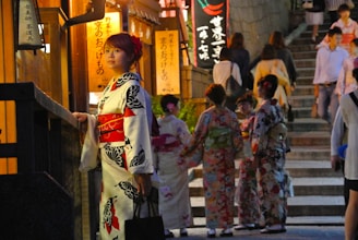 A woman in traditional Japanese attire stands beside a building, gazing thoughtfully. Behind her, a group of people dressed in colorful kimonos gather, possibly preparing for a cultural event. The setting appears to be a street with stone steps leading uphill, illuminated by warm lights and lanterns with Japanese writing.