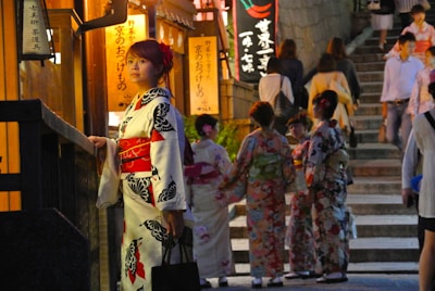 A woman in traditional Japanese attire stands beside a building, gazing thoughtfully. Behind her, a group of people dressed in colorful kimonos gather, possibly preparing for a cultural event. The setting appears to be a street with stone steps leading uphill, illuminated by warm lights and lanterns with Japanese writing.