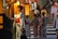 A woman in traditional Japanese attire stands beside a building, gazing thoughtfully. Behind her, a group of people dressed in colorful kimonos gather, possibly preparing for a cultural event. The setting appears to be a street with stone steps leading uphill, illuminated by warm lights and lanterns with Japanese writing.