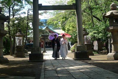 A couple wearing traditional Japanese attire walks through a path surrounded by stone lanterns and greenery. The woman holds a red parasol, and the man holds a purple parasol. The background features a traditional Japanese building with a green roof, partially obscured by trees.