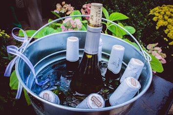 A metal bucket filled with several bottles of wine chilled in ice. The central bottle is prominently visible, with a cork slightly protruding. Around the bucket, various lush green plants and some pink and yellow flowers add a decorative touch.