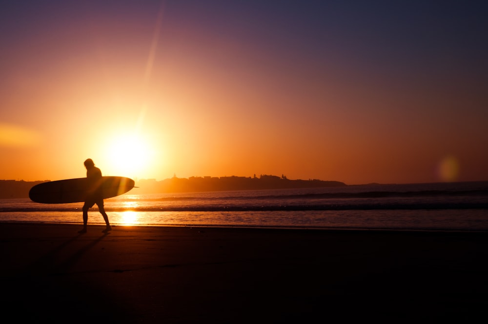 夕暮れ時に海岸近くでサーフボードを持つ男性のシルエット