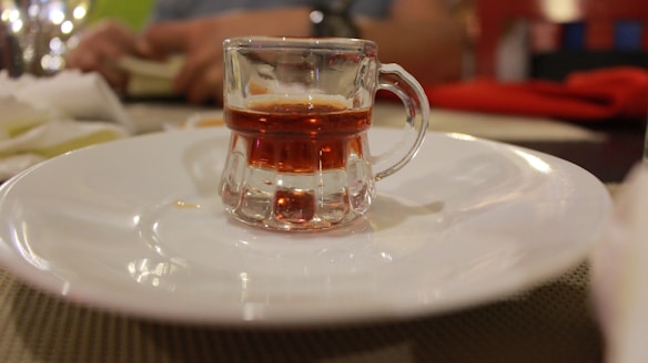 A small glass mug with a handle contains a dark amber liquid, placed at the center of a white plate. The background is blurred, featuring a person with a book and some brightly colored objects that appear to be napkins or place settings.