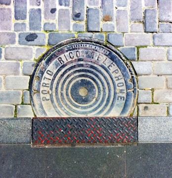 A round manhole cover on a pavement with the text 'Porto Rico Telephone' and circular patterns. It is surrounded by light gray stone bricks, and the lower section has a textured metal grate with red and black details. Small grass tufts are visible in the brick gaps.