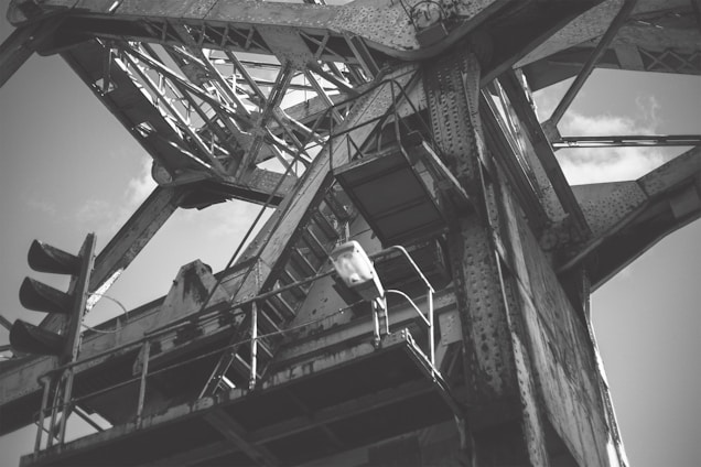 An industrial structure with metal beams and riveted joints, showcasing intricate latticework and towering framework against a partly cloudy sky.
