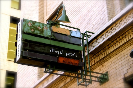 An outdoor restaurant sign with an artistic design featuring stars. The sign reads 'illegal pete's' and hangs from a historic-style brick building with ornate architectural details.