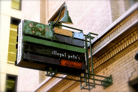 An outdoor restaurant sign with an artistic design featuring stars. The sign reads 'illegal pete's' and hangs from a historic-style brick building with ornate architectural details.