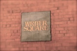 A brick wall with a metal plaque displaying the words 'WRITER SQUARE' in capital letters. The plaque has a rustic or vintage appearance, likely made of metal with an aged patina.