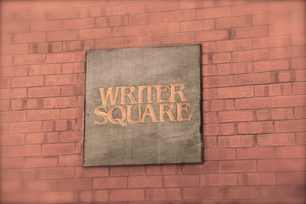A brick wall with a metal plaque displaying the words 'WRITER SQUARE' in capital letters. The plaque has a rustic or vintage appearance, likely made of metal with an aged patina.
