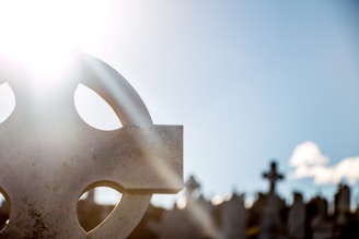 A bright sun shines over a cemetery, with a headstone in the foreground featuring a Celtic cross design. The background shows multiple other headstones silhouetted against a clear blue sky.