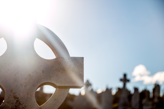 A bright sun shines over a cemetery, with a headstone in the foreground featuring a Celtic cross design. The background shows multiple other headstones silhouetted against a clear blue sky.