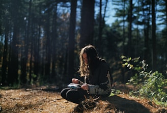 A person with long hair sits outdoors on a bed of dry leaves and pine needles in a forest. They appear to be engrossed in reading or writing, holding a small book or notebook. Sunlight filters through the tall trees, casting dappled light on the scene, and some green foliage is visible nearby.