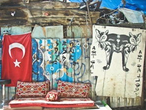 A rustic setup includes a weathered wooden wall adorned with a red flag featuring a white star and crescent. Beside it, vibrant, patterned rugs are placed on a bench, creating a cozy seating area. Nearby, a large artistic poster depicts a mirrored image of a cat, adding an element of street art.
