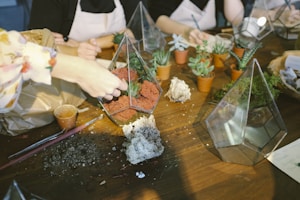 A group of people are assembling terrariums. On the table, there are geometric glass containers being filled with red soil and plants. Small potted succulents are scattered around, and there are tools like paintbrushes and tweezers. Hands are seen actively arranging materials in the containers.