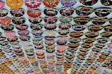Rows of intricately designed miniature hats with various vibrant colors and decorations are hanging on display. Each row features different patterns, including vibrant floral and geometric designs. The items are arranged neatly in a grid-like pattern against a light background.