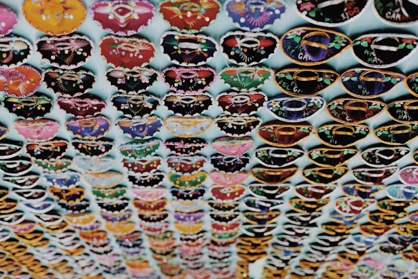 Rows of intricately designed miniature hats with various vibrant colors and decorations are hanging on display. Each row features different patterns, including vibrant floral and geometric designs. The items are arranged neatly in a grid-like pattern against a light background.