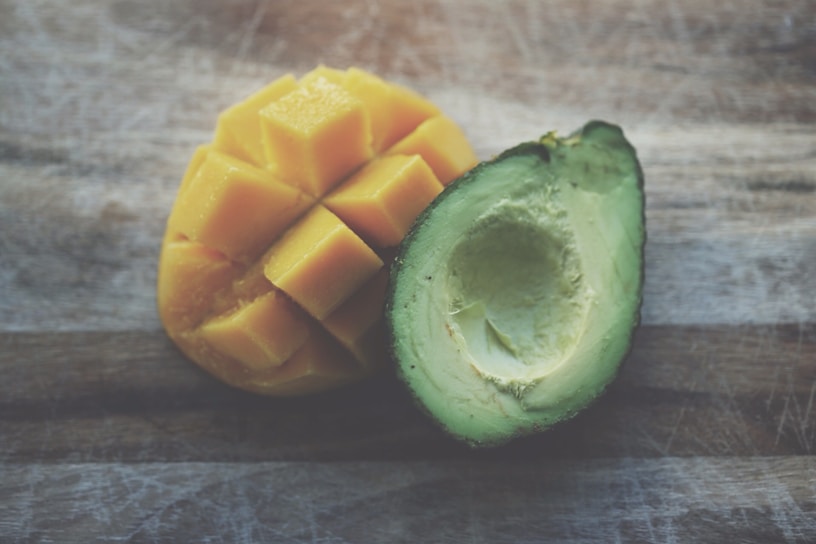 A halved avocado with a smooth green surface is placed next to a sliced mango, showcasing vibrant yellow cubes. Both fruits are on a textured wooden surface that contrasts with their colors.