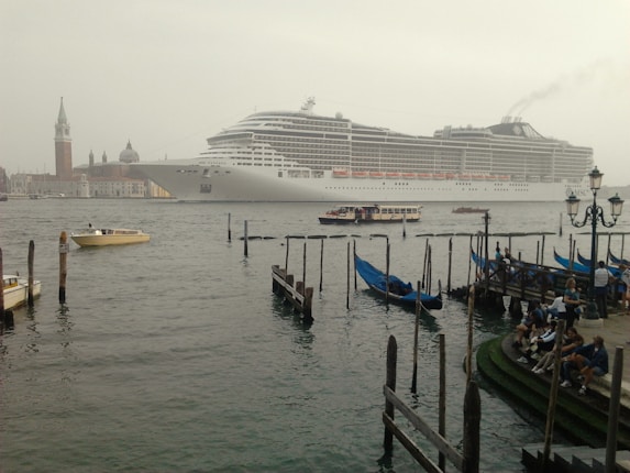 A large white cruise ship is sailing along a body of water, with smaller boats including gondolas and water taxis nearby. On the right, there is a dock with people sitting and observing the scene. In the background, historical buildings and a tall bell tower are visible.