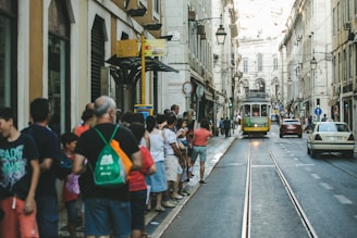 A street scene featuring a traditional yellow tram traveling down tracks lined with people on either side. The architecture includes tall, classic European-style buildings with arched windows and balconies. The atmosphere seems lively, with pedestrians standing close to the road, and a few cars are visible further down the street.