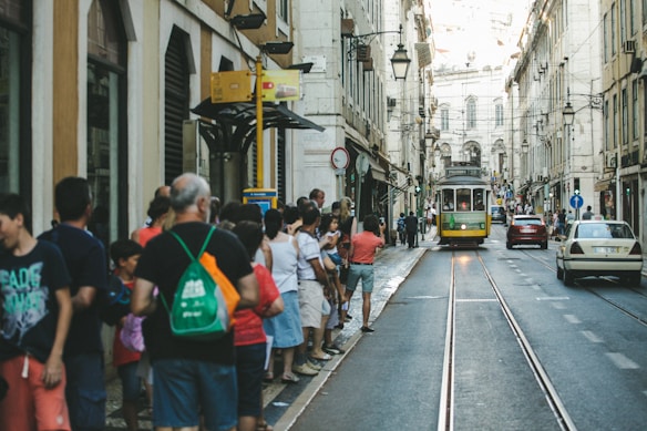 A street scene featuring a traditional yellow tram traveling down tracks lined with people on either side. The architecture includes tall, classic European-style buildings with arched windows and balconies. The atmosphere seems lively, with pedestrians standing close to the road, and a few cars are visible further down the street.