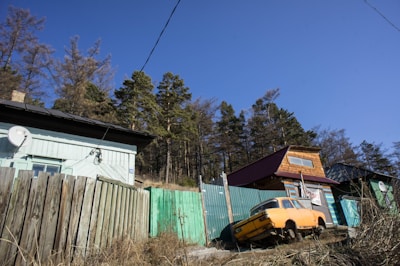 An image showcasing a rural setting with two wooden houses surrounded by a dilapidated fence. One house has a light blue color and a satellite dish, while the other has a mix of colors including turquoise and natural wood. An old, yellow car is partially visible, seemingly abandoned next to the fence. In the background, there is a forest with tall, sparse trees under a clear blue sky.