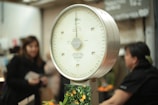 An analog scale with a white dial and a metallic frame is prominently featured. Numbers and markings in black are visible on the scale's dial. In the background, there are several blurred figures, possibly people, along with a blurry environment that suggests a market setting.