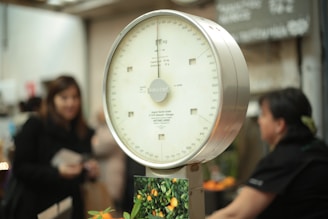 An analog scale with a white dial and a metallic frame is prominently featured. Numbers and markings in black are visible on the scale's dial. In the background, there are several blurred figures, possibly people, along with a blurry environment that suggests a market setting.