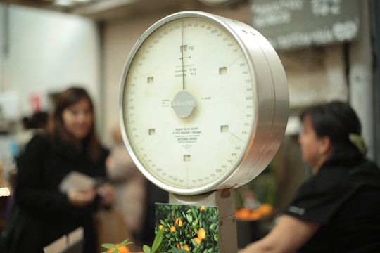 An analog scale with a white dial and a metallic frame is prominently featured. Numbers and markings in black are visible on the scale's dial. In the background, there are several blurred figures, possibly people, along with a blurry environment that suggests a market setting.