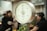 An analog scale with a white dial and a metallic frame is prominently featured. Numbers and markings in black are visible on the scale's dial. In the background, there are several blurred figures, possibly people, along with a blurry environment that suggests a market setting.