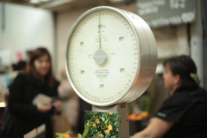 An analog scale with a white dial and a metallic frame is prominently featured. Numbers and markings in black are visible on the scale's dial. In the background, there are several blurred figures, possibly people, along with a blurry environment that suggests a market setting.