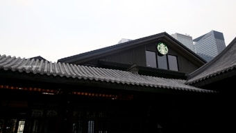 A traditional-style building with a tiled roof integrates a modern coffee shop, indicated by a green and white logo, set against a backdrop of modern skyscrapers.