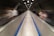 A long, straight moving walkway in an underground tunnel surrounded by metal walls. Blurred figures of people in motion are seen on both sides as they move along the walkway, contributing to a sense of speed and movement. Overhead lights illuminate the entire tunnel, creating a well-lit path.