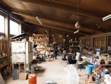 A spacious pottery studio filled with various pottery tools, materials, and equipment. The studio has large wooden shelves stocked with jars, pots, and other ceramic items. A kiln is present on the left side of the room, and numerous finished pieces are scattered throughout the area. Two people are engaged in their work amidst the cluttered yet creative environment, with wooden beams and large windows providing a rustic feel.