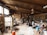 A spacious pottery studio filled with various pottery tools, materials, and equipment. The studio has large wooden shelves stocked with jars, pots, and other ceramic items. A kiln is present on the left side of the room, and numerous finished pieces are scattered throughout the area. Two people are engaged in their work amidst the cluttered yet creative environment, with wooden beams and large windows providing a rustic feel.