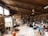 A spacious pottery studio filled with various pottery tools, materials, and equipment. The studio has large wooden shelves stocked with jars, pots, and other ceramic items. A kiln is present on the left side of the room, and numerous finished pieces are scattered throughout the area. Two people are engaged in their work amidst the cluttered yet creative environment, with wooden beams and large windows providing a rustic feel.
