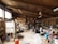 A spacious pottery studio filled with various pottery tools, materials, and equipment. The studio has large wooden shelves stocked with jars, pots, and other ceramic items. A kiln is present on the left side of the room, and numerous finished pieces are scattered throughout the area. Two people are engaged in their work amidst the cluttered yet creative environment, with wooden beams and large windows providing a rustic feel.