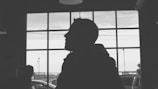 Silhouette of a person against a large window with a grid pattern. The scene is inside a building with two people partially visible, along with outdoor elements like vehicles and power lines visible through the window.