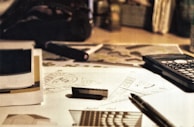 A workspace with technical drawings, a pen, a calculator, and a USB drive scattered on a desk. The setting suggests an environment focused on design or engineering, with blueprints and sketches partially visible. The lighting is warm and subdued.